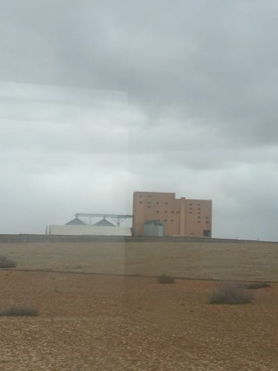 A beige building complex stands against a cloudy sky, featuring a tall, multi-story structure with several windows. Silos and industrial apparatuses are connected to it. A light brown, textured ground stretches to the right, marked by sparse vegetation. The overall scene evokes a sense of industrial or agricultural facility under an overcast sky. The photograph has been rotated.