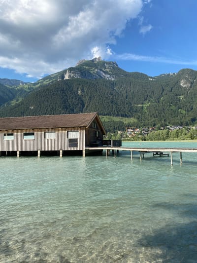 This is a photograph of a wooden boathouse situated on the turquoise waters of a lake in Lärchenwiese, Austria. The boathouse, constructed with vertical wooden planks, features several windows, some with fabric coverings. It is supported by stilts above the water and connected to a wooden pier that extends into the lake. The pier has wooden posts and a simple bench or platform. In the background, a dense forest covers the slopes of a large mountain, which is topped with rocky peaks. A small village is visible nestled on the far side of the lake, with buildings and some trees. The sky above is blue with scattered white and gray clouds. The water in the foreground is clear, allowing the lakebed to be partially visible, and exhibits ripples and reflections. A few people can be seen swimming or enjoying the water in the distance.