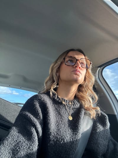 The scene depicts a female individual seated in the front passenger or driver's side of a vehicle, in what is stated as Chantemerle-les-Blés, France. No specific external landmarks of the city are visible within the frame. The individual, appearing to be a young adult, has wavy light brown hair. She wears large, tortoise-shell patterned, rectangular-framed glasses with a blue tint visible in the lenses. Her attire consists of a dark grey, knitted crew-neck sweater. She is accessorized with a gold-colored earring in her visible right ear and two gold-colored necklaces; one features a small circular pendant, and the other is a shorter, possibly beaded chain. A dark-colored seatbelt is fastened diagonally across her chest and left shoulder. The individual is looking upwards and to her right, exhibiting a neutral facial expression. Strong natural sunlight enters the vehicle from the right side, illuminating the right side of her face and hair, and creating prominent shadows. The vehicle's grey headliner is visible overhead. Through a portion of the vehicle's rear window on the left, a clear blue sky with some scattered white clouds is discernible.