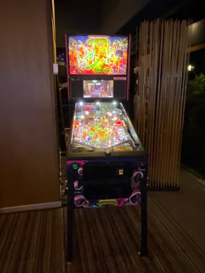 The image shows a Teenage Mutant Ninja Turtles pinball machine with a vibrant display in a dimly lit environment. The machine is positioned indoors, possibly in a game room or arcade. The background reveals a horizontally-oriented texture and a wooden panel to the foreground. The location is not directly discernible from the image.