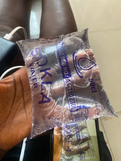 The image shows a close-up of a person's hand holding a clear plastic sachet of water labeled "SOKLA WATER." The sachet has blue text and a blue water droplet logo. Text on the sachet indicates a net volume of "50cl" and a registration number "NA1-110230L." Another identical sachet is visible in the background, partially obscured by the one in the foreground. The setting appears to be indoors, with a neutral-colored surface in the background. A portion of a black electronic device with a white cable is also visible in the lower left corner. The lighting suggests it could be daytime. The image focuses on the product, highlighting its branding and details.