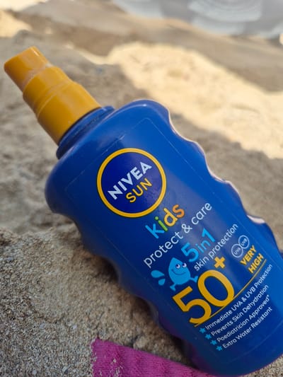 A blue bottle of Nivea Sun Kids 5-in-1 SPF 50 sunscreen lotion with a yellow spray nozzle lies on a sandy surface, possibly a beach. The bottle features the Nivea Sun logo with a yellow circle and the text "SUN" in yellow. Below that, it reads "kids protect & care" in various colors, and prominently displays "5in1 skin protection" with a cartoon water droplet character. A large yellow "50" is a prominent feature, along with smaller text indicating "UVA UVB VERY HIGH" and other benefits like "Immediate UVA & UVB Protection", "Prevents Skin Dehydration", "Paediatrician Approved", and "Extra Water Resistant". A small portion of a pink towel is visible in the bottom left corner. The background is out of focus, showing sandy mounds and sunlight casting shadows. The scene does not contain any people or indicate any specific city.