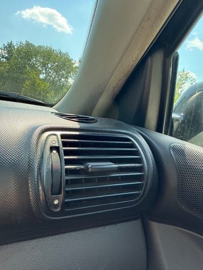 The image displays the interior of a vehicle, focusing on a central air vent and the surrounding dashboard. The vent has vertical slats and a circular dial below it for control. To the left of the vent, a portion of the windshield is visible, showing a bright blue sky with scattered white clouds and the green foliage of a tree. The dashboard material appears to be textured black plastic. In the upper left of the image, a black trim piece runs along the edge of what might be a sunroof or a window frame, also showing a glimpse of the sky. The lighting suggests it is daytime, likely late morning or early afternoon, with the sun casting shadows on the dashboard. There are no people visible. The scene is set within a car, suggesting the viewer is inside the vehicle. The overall impression is a close-up, internal view of a car's dashboard on a clear day, with the outside environment peeking through the windows.