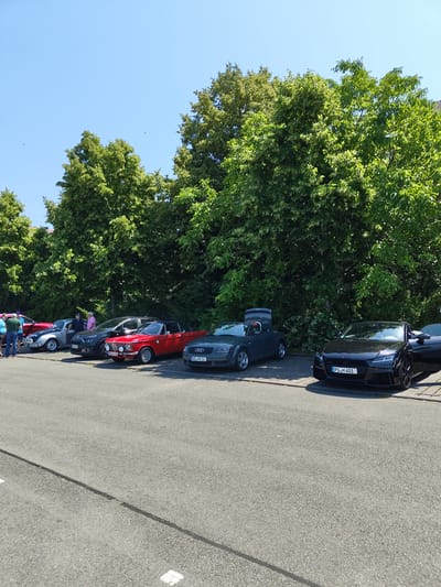 A row of parked cars is lined up on an asphalt surface in front of a dense line of green trees. The sky is clear and blue. Several people are standing near the cars on the left side of the image. The cars include a red convertible, a dark gray convertible with its trunk open, and a black sports car with its driver's side door open. The license plate on the black car reads "PS H 401". The scene appears to be outdoors in a park or parking lot.