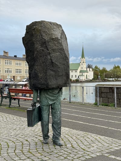 The image shows a statue in Reykjavik, Iceland. The statue depicts a man with a stone head sitting on a bench. The statue is dressed in a suit. Behind the statue is a building with many windows. To the right of the statue, across the street, is a waterway and a church with a green roof and spire. The street is paved with cobblestones.