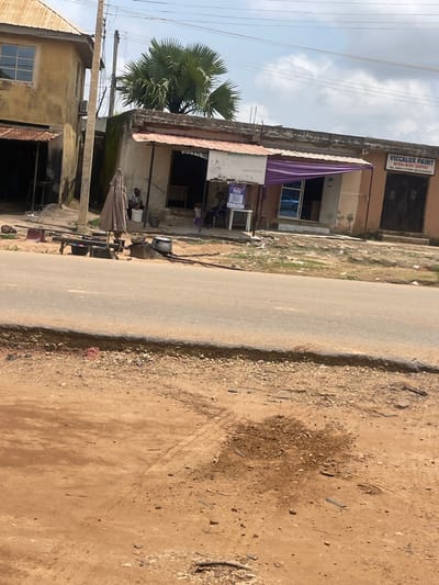 The image shows a street scene in Unguwar Saya, Nigeria. A two-story building with a weathered yellow facade and a corrugated metal roof is visible on the left, with a window on the upper floor and a covered entrance on the ground level. Adjacent to it, a single-story building has a similar corrugated metal roof. This building features a purple awning over an open storefront. To the right of the storefront, a sign reads "VICCALUX PAINT BYDA-BIDI DEPOT". There are dark wooden doors on the far right of this building.
In front of the buildings, a paved road runs horizontally across the image. On the side of the road, near the buildings, several objects are present. A person is seated on a low stool next to a white bucket. Nearby, there are cooking pots, a table with some items on it, and what appears to be laundry. A large umbrella is positioned over the table. Further to the right, two children are visible, one standing and the other seated.
The ground in front of the road is composed of reddish-brown earth with scattered stones and some vegetation. In the foreground, tire tracks are imprinted in the dirt. The sky above is partly cloudy. The overall scene depicts a typical roadside environment with commercial activity and residential elements.