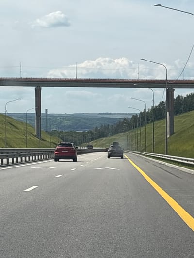 A highway overpass looms over a multi-lane asphalt road in an outdoor environment, likely in or near a city. The overpass is supported by two large concrete pillars. On the road, two cars are visible, one red and one dark-colored, traveling in the same direction. The red car is in the left lane, and the dark car is in the right lane, slightly ahead. The road is marked with white lane dividers and a yellow line down the center. The surrounding landscape features green, grassy hillsides and a line of trees in the distance. The sky is partially cloudy with a mix of blue sky and white clouds. Streetlights are positioned along the right side of the road. The image is taken from a vehicle's perspective, suggesting it was captured while driving. There are no people clearly visible in the image. The scene appears to be set in a relatively rural or suburban area, possibly during daylight hours given the brightness. The specific location within the city of Sapogoli, Russia, is not discernible from the visual cues alone.