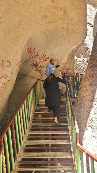 Three men are on a stairway. The wall to the side is off-white and has some graffiti. The stairs are painted with light green and brown. The person closest to the viewer is wearing a black dress and has bare feet. There is metal fencing along the side of the stairs.