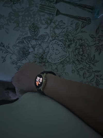 The image shows a person's arm with a smartwatch. The smartwatch has a round face with a black band. The screen of the watch displays the time as 22:28 and other glowing orange numbers. The person's arm is resting on a surface. In the background, there is a patterned tablecloth with floral designs and images of forks. The lighting suggests it is nighttime or indoors with dim lighting. The location is not specified in the image or the provided hints.