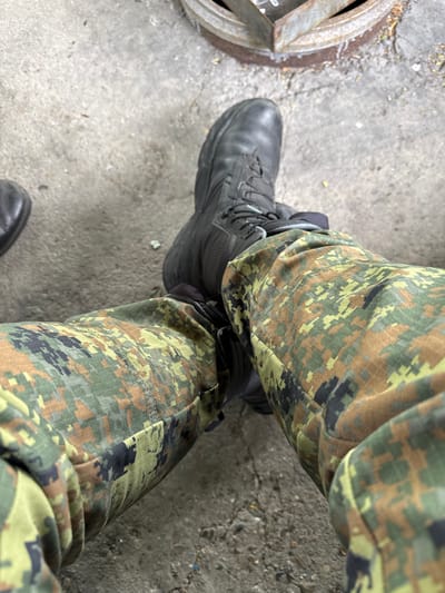 The image shows a person's legs and feet. They are wearing camouflage patterned pants and black leather boots. The person is standing on a concrete surface, near a metal object that appears to be part of a manhole cover. The image was taken in an outdoor location, potentially in a city such as Bozhurishte, Bulgaria.