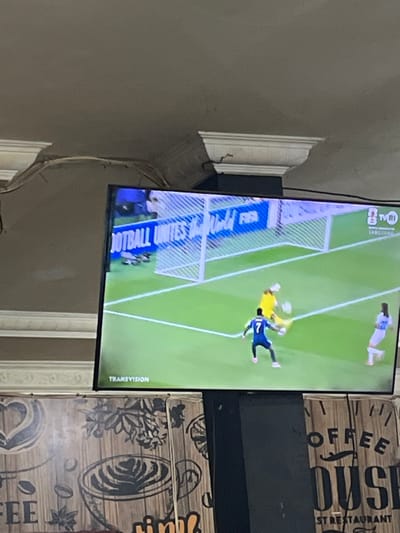 The image shows a television screen displaying a football match. A player in a blue uniform with the number 7 is attempting to score a goal, while the goalkeeper in yellow is attempting to save it. Another player in a white uniform is standing to the side. The background of the screen shows a blue banner with text that includes "FOOTBALL UNITES" and "FIFA". The TV is mounted on a black stand, and below the stand, there is a wooden surface with graphics that appear to be for a coffee house or restaurant, featuring images of coffee cups and text. The scene suggests the television is in a public place or a home setting where a sports event is being broadcast. The text on the wooden surface at the bottom indicates that this location is likely in Keudesampoiniet, Indonesia, due to the presence of the text "RST RESTAURANT".