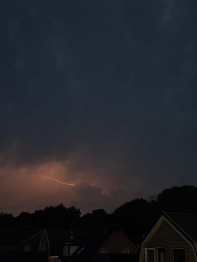 A lightning strike illuminates the dark, cloudy sky above the rooftops of homes in the city of Putten, Netherlands. The lightning appears as a bright, jagged line of light against a backdrop of deep blues and grays, with some orange hues bleeding from behind the clouds. Below the sky, silhouetted trees form a dark line across the middle of the image, separating the sky from the foreground. The tops of several houses are visible, with dark roofs and the outlines of windows and chimneys. The overall atmosphere is dramatic and captures a moment of a thunderstorm.