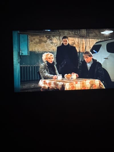 A medium shot shows three people gathered around a table in what appears to be an indoor, industrial-like setting. In the foreground, a woman with blonde hair and a leopard-print coat sits to the left of a checkered tablecloth. She holds a white mug and looks away from the camera. To her right, a man with blonde hair wearing a grey turtleneck and a dark coat sits at the table, also looking away. In the background, a third person, a man in a dark coat, stands behind the table. The environment features a radiator, a light fixture, and part of a white vehicle to the right. The overall lighting suggests an indoor scene, possibly a garage or workshop. There is no visual information within the image that indicates the location being the city of Skalyste, Ukraine.