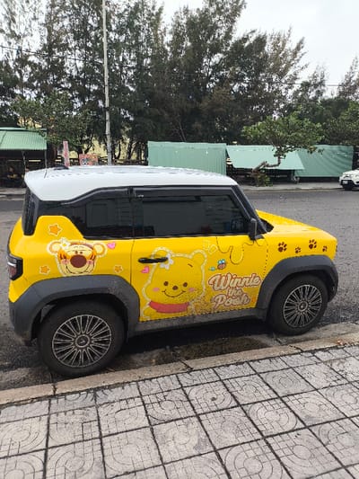 A yellow and white compact vehicle, adorned with "Winnie the Pooh" themed graphics including images of Winnie the Pooh and Tigger, honey drips, and paw prints, is parked curb-side on a street in Thành phố Tuy Hòa, Vietnam. The vehicle features a white roof, yellow body, black window trim, black door handles, and black fender flares over multi-spoke alloy wheels. The car is positioned on asphalt pavement, adjacent to a grey sidewalk composed of patterned square tiles.
In the background, a row of tall, slender evergreen trees dominates the skyline, suggesting a coastal or park environment. Beneath the trees, several structures are visible. To the left, a green-roofed open-air stall or small building contains indistinct figures. To the right and center, multiple structures made of light green corrugated metal are present, possibly temporary shelters or vendor stalls. A tall utility pole rises in the mid-ground. A white car is partially visible parked on the far right of the street in the distance. The sky is overcast. No overt human actions or interactions are prominently displayed in the immediate foreground.