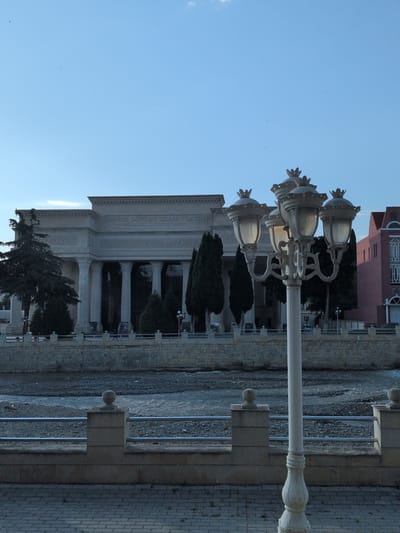 The image depicts the Ganja State Drama Theater in Ganja, Azerbaijan, a neoclassical building with grand columns and an inscription above the entrance. A decorative lamppost with four lights stands in the foreground, its ornate design contrasting with the solid architecture of the theater. The theater is situated across a dry riverbed lined with stone walls and metal railings. Several dark, conical trees are planted in front of the building. To the right of the theater, a pink building with arched windows is partially visible. The sky above is a clear, pale blue with a few faint clouds and distant birds. The ground in the foreground is paved with interlocking bricks, suggesting a public walkway or plaza.
