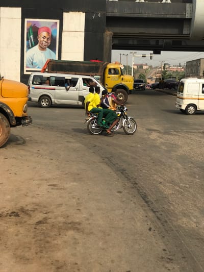 The scene depicts a busy road junction or segment under an overpass in Umuokpu, Nigeria, during daylight hours. The road surface is paved and shows significant wear, including dirt, tire marks, and oil stains.
In the mid-ground, three individuals are riding a silver motorcycle from left to right. The primary rider wears an orange shirt. The two passengers behind him wear matching yellow shirts and dark green trousers, indicative of school uniforms.
Vehicular traffic is present. To the left, a partial view of a yellow truck's front fender and wheel is visible. Behind the motorcycle, a silver commercial van (likely a Toyota Hiace) is present, with several occupants visible through its windows, one looking out. Behind this van and further right is a yellow dump truck with a dark, open bed and a Mercedes-Benz grille. To the far right, a white commercial panel van, marked "TRANSPORT COMPANY OF NIGERIA" (partially visible), is also present. Several other vehicles are visible in the background, ascending a hill.
An overhead bridge or overpass spans the road. Its supporting columns are painted with prominent black and white vertical stripes. On the left column, a large portrait poster depicts a dark-skinned man smiling, wearing a light-blue traditional tunic and a red cap, against a background featuring green, white, and black stripes. Traffic lights are visible above the road under the overpass. The background reveals a residential or semi-urban area with scattered buildings and utility poles under a hazy sky.