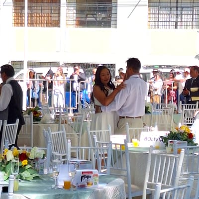 A wedding reception unfolds in Cerro Colorado, Peru, featuring a couple in a close embrace. A light-skinned woman with dark hair, adorned in a white dress, is embraced by a light-skinned man wearing a white shirt. They stand amidst rows of white chairs set up for the celebration.

Attendees of the reception are visible in the background, seated at tables. The tables are decorated with flowers. A sign reading "Bolivia" is visible. The setting appears to be partially outdoors, with a building featuring barred windows situated nearby.