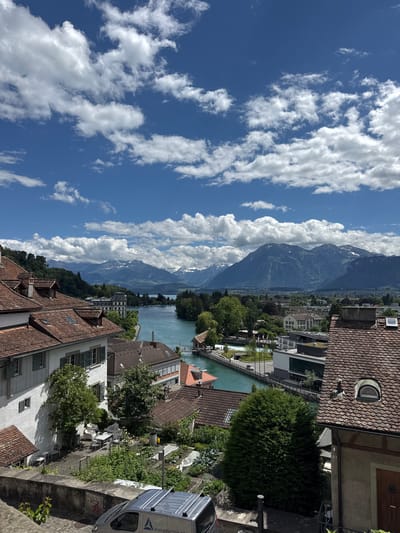 The image depicts a scenic view of the city of Thun, Switzerland, focusing on its charming architecture, the turquoise waters of the Aare River, and the majestic Swiss Alps in the background. The foreground is dominated by traditional Swiss-style buildings with red tile roofs, white facades, and dark green shutters. One prominent building features an outdoor seating area with white furniture. A small garden with various plants is visible next to it. A silver van with the text "ponta energie technik.ch" is parked on a cobblestone street in the lower left portion of the frame.

The middle ground showcases the Aare River, its vibrant blue-green color contrasting with the surrounding greenery and buildings. Several trees line the riverbanks, and a pedestrian bridge crosses over the water. Modern buildings with dark roofs are interspersed with older structures along the river. In the distance, a large body of water, likely Lake Thun, is visible, leading towards the imposing silhouette of the snow-capped Alps. The sky is a bright blue, adorned with fluffy white cumulus clouds, suggesting a clear and pleasant day. The overall scene conveys a sense of tranquility and natural beauty characteristic of Swiss landscapes.