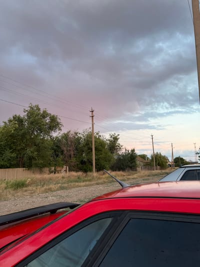 The image shows a rural street scene in Rakhinka, Russia, with a dramatic sky. In the foreground, the top and side of a red car are visible, its surface showing water droplets. Behind the car, dry, tall grass lines a dirt road. A wooden fence runs along the edge of the property, with dense green trees and some dry brush behind it. Several utility poles with power lines crisscross the sky, which is filled with heavy, gray clouds tinged with pink and pale blue from the setting sun. In the middle ground, houses with wooden fences are visible amongst the trees. Another car, silver or white, is parked further down the road. The overall impression is one of a quiet, possibly impending storm in a rural, residential area.