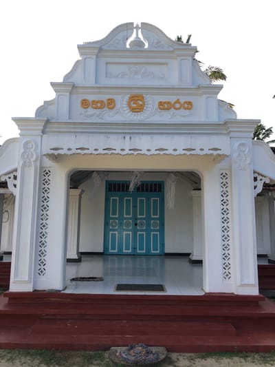 This is a frontal, eye-level, outdoor shot of the entrance to a white building with ornate architectural details, located in Pilana, Sri Lanka. The building features a decorative pediment with yellow lettering, flanked by pillars with intricate carvings. A light blue double door with white trim is visible beyond an archway, which is decorated with white paper streamers. White scalloped trim runs along the underside of the archway. The entrance is accessed by a set of wide, red-painted steps. The ground in front of the steps is covered in patchy grass. The sky is bright and overcast.