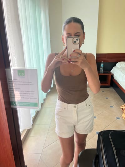 A female subject is captured in a mirror selfie within a hotel room located in Chamilpa, Mexico. She is positioned facing a large, framed mirror, holding a light-colored smartphone with both hands to obscure her face while capturing her reflection. The subject wears a brown square-neck tank top and white cuffed shorts. Adornments include gold-toned hoop earrings, a delicate necklace with a circular pendant, and rings on her left ring finger and right middle finger; her fingernails are painted red.

The reflected environment reveals a light yellow wall and a tiled floor with a pattern of light beige squares accented by smaller dark blue squares. To the left, a window is covered by light green sheer curtains. To the right, a bed with white linens and a dark brown headboard is visible, alongside a dark wooden nightstand holding a plastic water bottle. A black piece of luggage is partially visible on the floor in the bottom right. A laminated sign with "GS Hoteles" branding and Spanish text regarding water conservation and towel changing policies is affixed to the left side of the mirror or an adjacent surface.