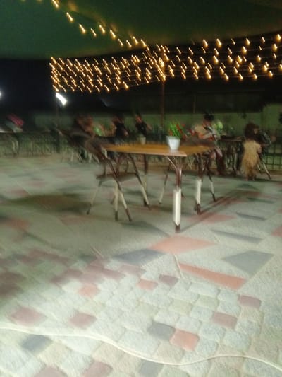 This image depicts an outdoor seating area at night, likely a cafe or restaurant, with decorative string lights overhead casting a warm glow. The setting appears to be located in Dustlik, Uzbekistan, suggested by the context. In the foreground, a patterned tiled floor is visible, with a slight blur due to motion. In the mid-ground, several tables and chairs are arranged, some occupied by blurred figures. One table in the center features a white pot with greenery and red flowers, adding a touch of color to the scene. The overall ambiance is relaxed and inviting, characteristic of an evening gathering. The lighting suggests it is nighttime, and the absence of harsh shadows implies mild weather conditions. The people in the image are indistinct due to the blur, making it difficult to discern specific details about their activities or emotions, though they seem to be seated and engaged in conversation or dining. No visible text is present in the image.