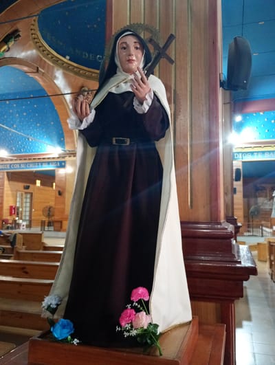 The image depicts a statue of a Catholic saint, likely Teresa of Avila, positioned inside a church. The location is the city of Pozo Almonte, Chile. The statue is a figure of a woman in a brown habit, white wimple and cape, holding a crucifix in her left hand and extending her right hand in a gesture of blessing. The statue is placed on a wooden stand adorned with colorful artificial flowers. The church's interior is visible in the background, featuring wooden structures, including pews and columns, and a blue ceiling decorated with star-like patterns. A loudspeaker is mounted on one of the columns, and religious inscriptions are present on the walls. The scene is bathed in ambient light, creating a somber yet reverential atmosphere.