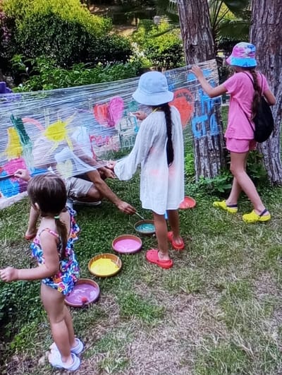 The image captures an outdoor art activity in Kichik Tasmachi, Uzbekistan, involving four individuals painting on a transparent film stretched horizontally in a grassy area bordered by dense foliage and trees.
In the foreground, a young female child with dark hair in pigtails wears a multicolored swimsuit and white Crocs-style sandals, standing with her back partially to the viewer, facing the transparent film. To her right and slightly behind, an adult, only partially visible from the waist down, wears light-colored shorts and dark footwear, crouching or kneeling near the lower section of the transparent film.
To the right of the adult, a female child in a light blue bucket hat, a white long-sleeved tunic, and red Crocs-style sandals actively applies paint to the transparent film with a brush. Her dark hair is in a single braid. Further to the right, another female child, wearing a multicolored bucket hat, a pink t-shirt, pink shorts, and bright yellow Crocs-style sandals, also paints on the film. She carries a black backpack on her left shoulder.
The transparent film displays various colorful painted marks, including hearts and abstract designs in red, pink, yellow, green, and blue. Five shallow, round bowls containing liquid paints in shades of turquoise, pink, yellow, and purple are arranged on the grass directly in front of the participants. The ground is covered in green grass with some visible soil patches. The background features lush green vegetation, including tree trunks on the right side of the frame.