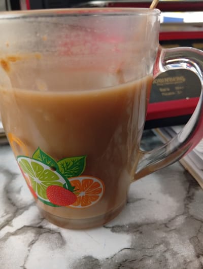 A close-up shot shows a partially filled glass mug decorated with images of sliced citrus fruits (lime and orange) and a strawberry. The mug is on a surface with a marbled pattern. The contents of the mug appear to be a beige-colored liquid, possibly coffee or tea with milk. Part of a red object with text is visible in the background. The location and city cannot be determined from the image.