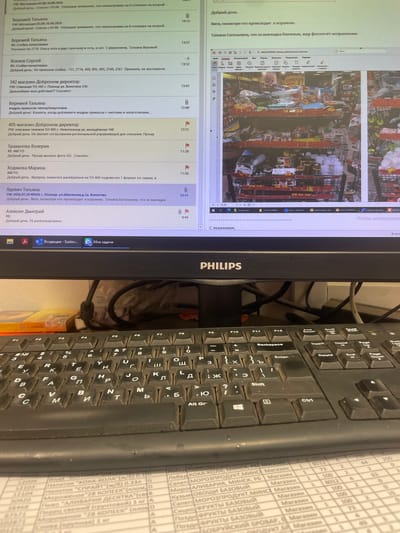The image shows a computer monitor displaying an email client with a list of messages and a web browser open to an online store, likely within a retail setting. The email client lists several messages with sender names like "Veremey Tatyana," "Hohlov Sergey," and "Trakhanova Valeria," and the content suggests work-related communication regarding merchandise, store operations, and inventory. One email specifically mentions "405 magazin Dobronom director" and a request for a regional coordinating manager's approval for a write-off. Another email from "Yadyevich Tatyana" details a location in "p. Polotsk, ul. Shkolnaya, d.1a, Konechka," indicating a possible address within the Voropaevskiy selskiy Sovet, Belarus.

The web browser displays a visual of store shelves stocked with various goods, including canned items, packaged foods, and beverages. A shopping cart is partially visible, filled with merchandise. The overall environment appears to be an office or backroom area within a retail store, with a keyboard and monitor in the foreground, suggesting the user is actively working. The scene is lit by artificial light.