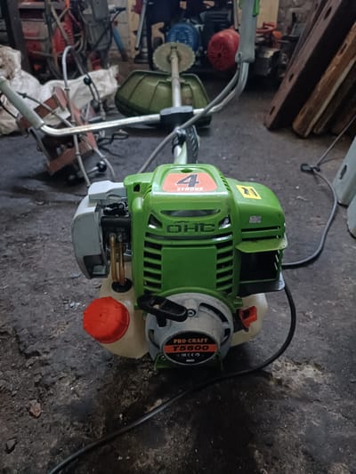 An outdoor, close-up, medium shot captures a green and grey gas-powered string trimmer engine resting on a dirt and asphalt surface. The engine, which appears to be a "PRO-CRAFT T5600 4-STROKE OHC" model, is the primary subject, positioned slightly off-center to the right. Its bright green casing is prominent, with a silver cylinder visible on the left side and an orange fuel cap on the bottom left. A black power cord snakes away from the engine towards the right edge of the frame. In the background, various other tools and machinery are visible, suggesting a workshop or storage area. The overall scene is somewhat cluttered, with debris and other objects scattered on the ground. This image was taken in Vuhlehirsk, Ukraine.
