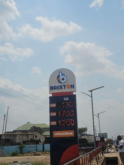 The image presents an outdoor daylight scene in AlakIa, Nigeria, centered on a tall, black and orange digital price sign for a 'BRIXTON' fuel station. The sign displays fuel prices: 'PMS 1730', 'AGO 1700', and 'LPG 1200', with 'WELCOME' illuminated at its base. A white arched panel at the top features the 'BRIXTON' name and a stylized 'b' logo with an orange teardrop. The sky above is clear blue with scattered white cumulus clouds. To the left of the sign, utility poles with power lines are visible, alongside a distant building with a green pyramidal roof, a lower building behind a light blue wall, and a pile of construction aggregate on the ground. To the right, multiple streetlights equipped with solar panels extend along a thoroughfare. A brown metal railing runs parallel to a drainage culvert in the mid-ground. On the far right, a male individual in a white shirt is partially visible from the back, along with other figures further in the background and partial signage for a 'PHARMACY' and an 'IN' entrance.