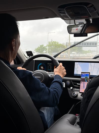 Here is a description of the image:

The image captures the interior of a car, with a focus on the driver. The driver is wearing a dark blue sweater and light blue jeans, with their hands on the steering wheel. The dashboard and infotainment system are visible, displaying information such as speed (96 km/h), driving statistics, and navigation details in Korean. A smartphone is mounted next to the infotainment screen, also showing navigation information. Outside the car, the windshield shows rain, and the view includes trees, streetlights, and other vehicles, indicating urban surroundings.

Based on the navigation display, the car seems to be in Goyang-si, South Korea, navigating towards a destination "679m 김포 (Gimpo)," with an alternative route available at "364m." The car appears to be in motion, as suggested by the speed displayed on the dashboard.