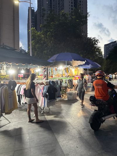 The image depicts a street scene at dusk in Haizhu District, China. A woman with long dark hair is browsing clothing racks at an outdoor market. She is wearing a black t-shirt and white shorts. In the background, a woman in a grey dress walks past a stall displaying various garments. To the right, a person on a black scooter wears an orange jacket and a red helmet. The scooter has a license plate reading "EG6468". Overhead, a blue umbrella covers some of the stalls. The ground is paved with grey tiles. In the distance, tall buildings rise against a twilight sky with scattered clouds. A street lamp illuminates the area on the left.