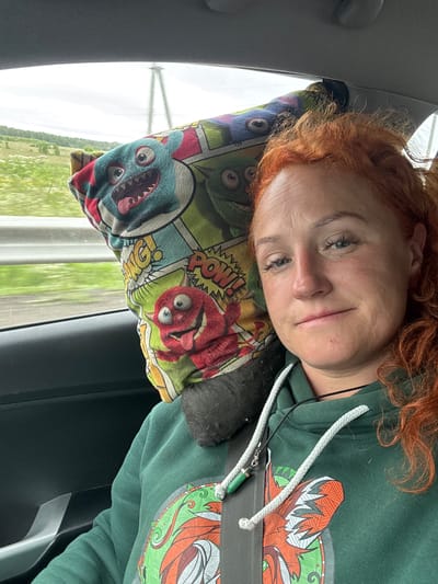 A woman with red hair, wearing a green hoodie and a seatbelt, is in the passenger seat of a car. A cartoon-themed pillow with monster illustrations is behind her head. The car is in motion, as indicated by the blurred background of trees and fields. The weather appears overcast. The location is not identifiable as being in the city of Можга, Russia.