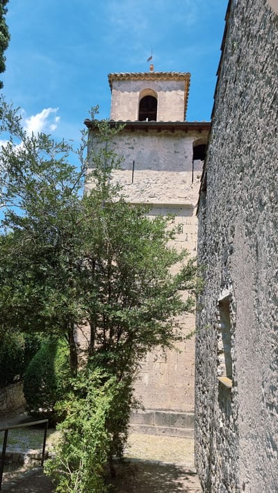 The image depicts an outdoor scene featuring an old stone structure, possibly a church or a watchtower, under a clear blue sky with scattered white clouds. The building has a weathered appearance, with rough-hewn stone blocks forming its walls and a terracotta-tiled roof. A prominent feature is a bell tower with arched openings, partially obscured by a lush green tree in the foreground. The tree's branches are dense with leaves, casting dappled shadows on the stone ground. The sun appears to be shining, suggesting it's daytime, likely late morning or afternoon, given the bright lighting and shadows. The overall atmosphere is serene and somewhat rustic, evoking a sense of history and tranquility. The location is identified as Sales de Llierca, Spain. There is no visible text in the image.