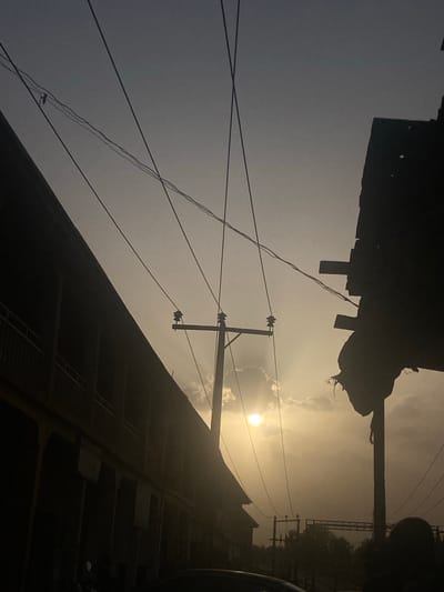 The image is an upward-angled shot of a street scene in Unguwar Fatika, Nigeria. The sun is positioned near the center of the frame, partially obscured by clouds, with rays of light breaking through. Power lines run diagonally across the sky, attached to utility poles. Silhouetted buildings are visible along the bottom right edge of the frame, with what appears to be scaffolding or a building facade featuring repetitive rectangular patterns. In the upper left corner, there is a darker, undefined object, potentially a building overhang or another structure partially obscuring the sky. The overall lighting creates a stark contrast between the bright sky and the dark silhouettes of the urban environment.