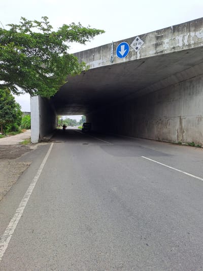 The image shows the entrance to a concrete underpass on a road in Cottunong, Indonesia. A tree with green foliage extends from the left side of the image, partially obscuring the top of the underpass. A traffic sign is visible on the right side of the underpass, indicating a height restriction of 4.8 meters. A blue circular sign with a white downward-pointing arrow is to the left of the height restriction sign. Inside the dimly lit underpass, a person on a motorcycle is visible in the distance, followed by a dark-colored vehicle. The road surface is asphalt with a white line marking the lane. The surrounding environment appears to be rural or semi-urban, with vegetation visible on the left side of the road. The sky is overcast.