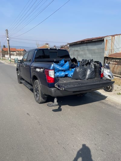 This image depicts a dark blue Nissan Titan PRO-4X crew cab pickup truck parked on the right side of an asphalt road in Resilovo, Bulgaria, under a clear, light blue sky. The truck is oriented facing away from the viewer, with its tailgate lowered. The truck bed is filled with waste bags: several blue bags are clustered on the driver's side, and multiple black bags are on the passenger's side. A black, retractable step is extended from beneath the truck's rear bumper on the driver's side.
On the left side of the road, a utility pole with multiple overhead power lines is visible, extending across the frame. Beyond the road and utility pole, residential buildings with red-tiled roofs and white facades are visible, set against distant hills or mountains. The ground on the far left shows sparse vegetation.
On the right side of the truck, a weathered building with corrugated metal siding and a rusty metal roof is partially visible. A silver-colored scooter or moped is parked to the right of the truck's rear, with its front portion obscured by the truck bed. A wire fence is visible further to the right, next to a patch of dirt and sparse grass. The asphalt road in the foreground shows minor markings and a distinct human shadow, indicative of the photographer, cast towards the lower left. No individuals are otherwise visible within the frame.