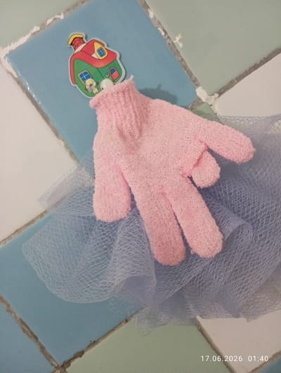 A pink, textured exfoliating glove is draped over a light blue mesh fabric. The glove is positioned with its fingers extended. Behind and slightly above the glove, a small, colorful sticker depicting a house with a dog is attached to a blue tile. The background consists of a tiled surface, with diagonal grout lines visible between the tiles, which are colored light blue, pale green, and white. The timestamp "17.06.2026 01:40" is present in the lower right corner. The scene appears to be indoors, possibly in a bathroom, given the tiled surface and exfoliating glove. The location is identified as Ohangaron, Uzbekistan.