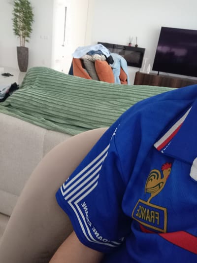 A person is sitting on a beige couch, wearing a blue France national football team jersey. A green ribbed blanket is draped over the couch. In the background, a television is visible, and a potted plant sits on a shelf. The location is inside a residence, most likely in Felgueiras, Portugal. No interactions or specific actions are taking place.