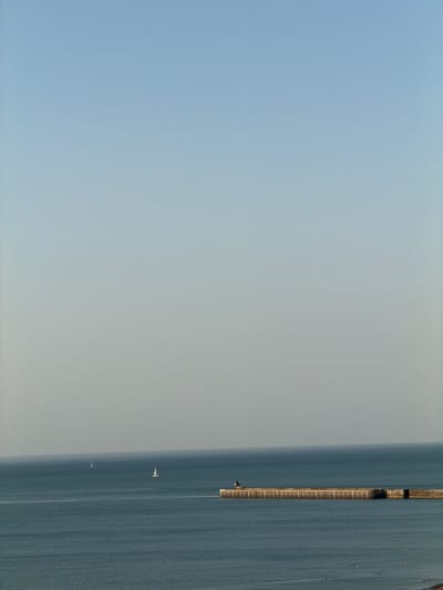 This is an aerial, wide shot of a calm, blue sea with a long pier extending into the water from the right side of the frame. The pier has a weathered, wooden surface and appears to be a popular spot for recreation, with several small yellow buoys floating in the water near it. In the distance, two sailboats are visible on the water, one larger and closer than the other. The sky is a pale, hazy blue, suggesting it might be early morning or late afternoon, with the sun not directly visible. The overall atmosphere is peaceful and serene. There are no people or visible text in the image. The image was taken in Dieppe, France.