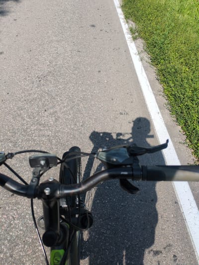 The image shows a close-up view of a bicycle's handlebars and part of the front wheel, from the rider's perspective. The handlebars are black and feature a gear shifter and brake lever. Cables for the brakes and gears run down from the handlebars. The bicycle is on an asphalt road, marked with a white line on the right side, separating it from a patch of green grass and low-lying vegetation. The sun is casting a shadow of the bicycle, including the rider, onto the road, indicating it is daytime and likely sunny. The scene appears to be outdoors in a location that has both paved surfaces and natural greenery, possibly a road or bike path in a rural or suburban area. There is no visible text in the image.