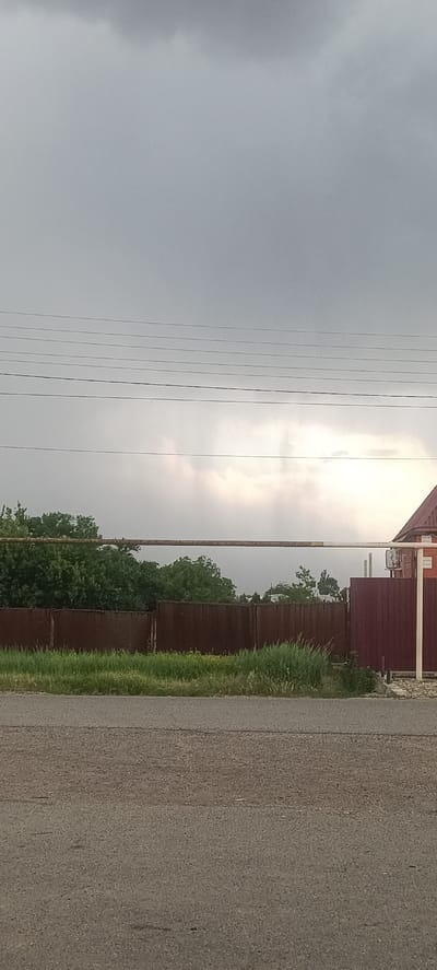 This is a vertical, medium-shot, outdoor photograph taken in Ivanovskaya, Russia. The image depicts a cloudy, overcast sky with what appears to be distant rainfall or a heavy fog obscuring the horizon. Several power lines are visible stretched horizontally across the upper portion of the frame.

In the middle ground, a rusty metal pipe runs horizontally, supported by posts. Behind this pipe, a dense line of green trees forms a backdrop. In front of the trees and the pipe, a weathered, dark brown metal fence runs across the majority of the image. To the right of the fence, a portion of a house with a red roof and brick exterior is visible.

The foreground is dominated by a grey asphalt road. The overall lighting suggests it is daytime, but the heavy cloud cover creates a subdued, potentially melancholic atmosphere. The scene appears to be rural or suburban, with the presence of houses, fences, and trees. There is no visible text in the image.