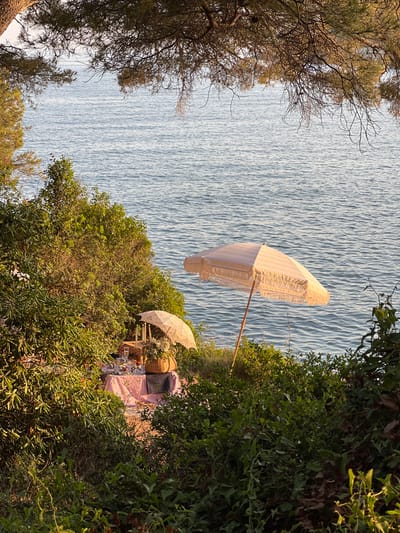 A serene seascape dominates the scene, bordered by foliage. A large, striped parasol leans toward the water, casting shade over an outdoor setting. A smaller parasol covers a picnic arrangement with pillows and wicker baskets. The scene exudes a calm, coastal vibe, with the lush greenery contrasting with the sparkling water.