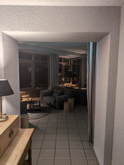 This image captures an interior view of a room, likely a living area, seen through a doorway. The room is furnished with a dark-colored sofa, a small wooden table with two glasses and what appears to be an orange drink, and two cylindrical wooden stools in the foreground. A dark lampshade is visible on the left side of the frame. The floor is tiled in a neutral gray. Large windows dominate the back wall, offering a view of a dark outdoor space, suggesting it is nighttime. The architecture features textured walls and a prominent doorway framing the scene. Based on the provided context, this scene is located in St. Märgen, Germany.