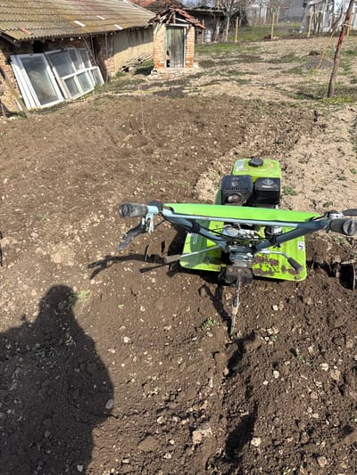 This forensic description details an outdoor scene in Profesor Ishirkovo, Bulgaria. The image, taken under bright daylight, shows a partially cultivated garden plot. In the foreground and mid-ground, dark brown, freshly tilled soil is prominent, exhibiting an uneven, clumpy texture. A bright green and black rototiller, featuring a visible engine block and handlebars, is positioned centrally in the mid-foreground, its tines partially submerged in the tilled earth, indicating a pause in cultivation. A distinct, elongated human shadow, consistent with a person holding a camera or phone, is cast across the lower left portion of the tilled ground. In the background, a single-story building with a tiled roof is visible on the left, against which several old white-framed window sashes are propped vertically. To its right stands a small, dilapidated brick shed with a wooden door and a sloped roof. Further into the background, sparse dry vegetation and additional distant structures can be discerned. A thin wooden stake protrudes from the tilled soil in the mid-ground, and a taller stake with a yellow top is visible further back on the right, accompanied by a thin yellow line or cable.