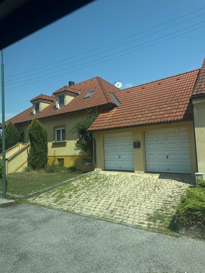 The image shows a residential property with a yellow house featuring a red tile roof under a clear blue sky. The house has dormer windows and a satellite dish on the roof. In front of the house, there is a double garage with two white sectional garage doors. The driveway is paved with light-colored interlocking stones, with patches of grass growing between them. To the left of the garage, a set of yellow stairs leads up to the house. Several green trees and bushes are present on the property. The scene appears to be in Wolfsthal, Austria, based on the provided context.