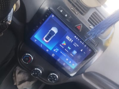The image shows the interior of a car, focusing on the dashboard and infotainment system. The car appears to be parked or in a stationary position. A portion of a steering wheel, covered with a patterned material, is visible on the left side of the frame.

The central element is a large, touch-screen infotainment display, which is on and showing various icons and information. The display shows a digital clock reading "06:51", a stylized car icon, and icons for applications such as Google Play and a music player. There are also navigation arrows and other status indicators. The screen's interface suggests it is running an Android-based system.

Below the infotainment screen, there are several physical controls. Two rotary knobs with metallic finishes are visible. The upper knob has red and blue indicators, likely for temperature control, and a central button. The lower knob appears to have multiple functions indicated by icons.

To the right of the infotainment screen, there are several buttons. One is a hazard light button with a red triangle symbol. Above that, there are two lock buttons. An air vent is also visible.

A blue fringed object, possibly a car accessory or decoration, is draped over the dashboard and partially obscures the air vent and infotainment screen. The interior of the car appears somewhat worn or dusty, with some reflections on the surfaces.

The image does not contain any explicit information that would pinpoint its location in Margabat, Uzbekistan. The visual elements are typical of a car interior and do not include any identifiable landmarks or context specific to that city.