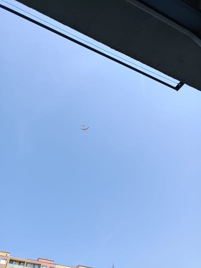 The image shows a clear, blue sky with a single airplane in flight. The airplane is white and appears to be a passenger jet, seen from below and slightly to the side. A portion of a building with multiple floors and balconies is visible at the bottom of the frame, suggesting an urban setting. The top of the image features the underside of an overhang or balcony, casting a shadow. The scene is likely from the city of Košice, Slovakia, given the presence of characteristic apartment buildings and the overall environment.